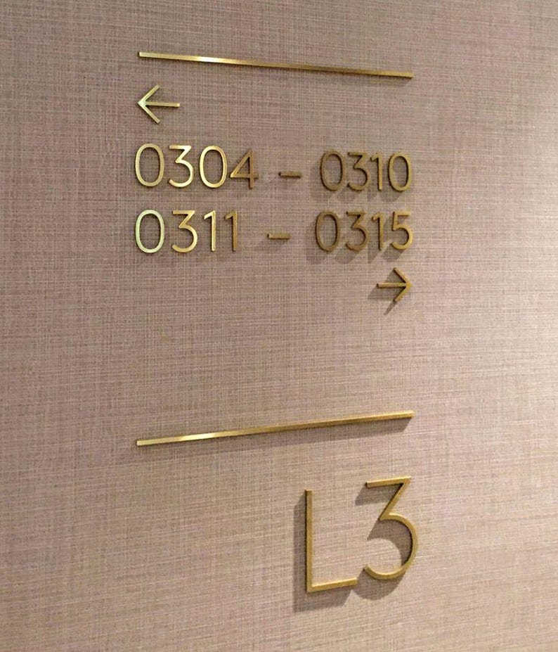 K&ouml;nnte beinhalten: Goldfarbene Richtungs- und Zimmernummernschilder an einer strukturierten, hellbraunen Wand. Das Schild enth&auml;lt Pfeile, Zimmernummern 0304-0310, 0311-0315 und die Etagenbezeichnung L3.