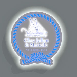 May include: A white circle with a blue rope border and a white sailboat illustration inside. The text "Chez Julien & Marielle POISSONNERIE" is written in black below the sailboat.