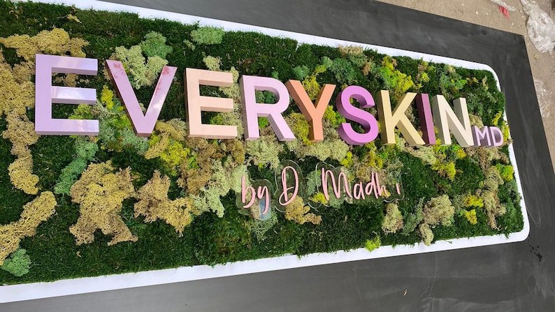 May include: A rectangular wall sign featuring a background of preserved green moss in various shades. Mounted on the moss are three-dimensional letters spelling EVERYSKIN MD in pastel colors, with a clear acrylic sign below reading by D Madhi.