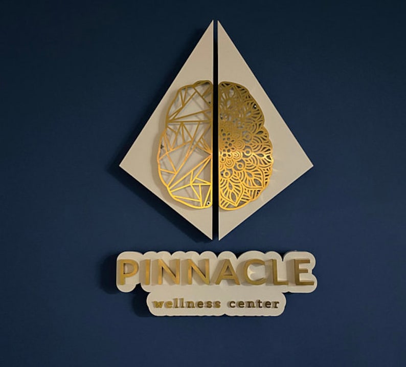 Puede incluir: Un letrero blanco en forma de diamante con un dise&ntilde;o de cerebro dorado en el centro. El letrero dice "PINNACLE wellness center" en letras doradas.