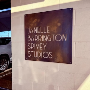 May include: A square, weathered metal sign with the text "JANELLE BARRINGTON SPIVEY STUDIOS" cut out. The sign is mounted on a white brick wall. The sign has a dark brown, rust-like appearance.