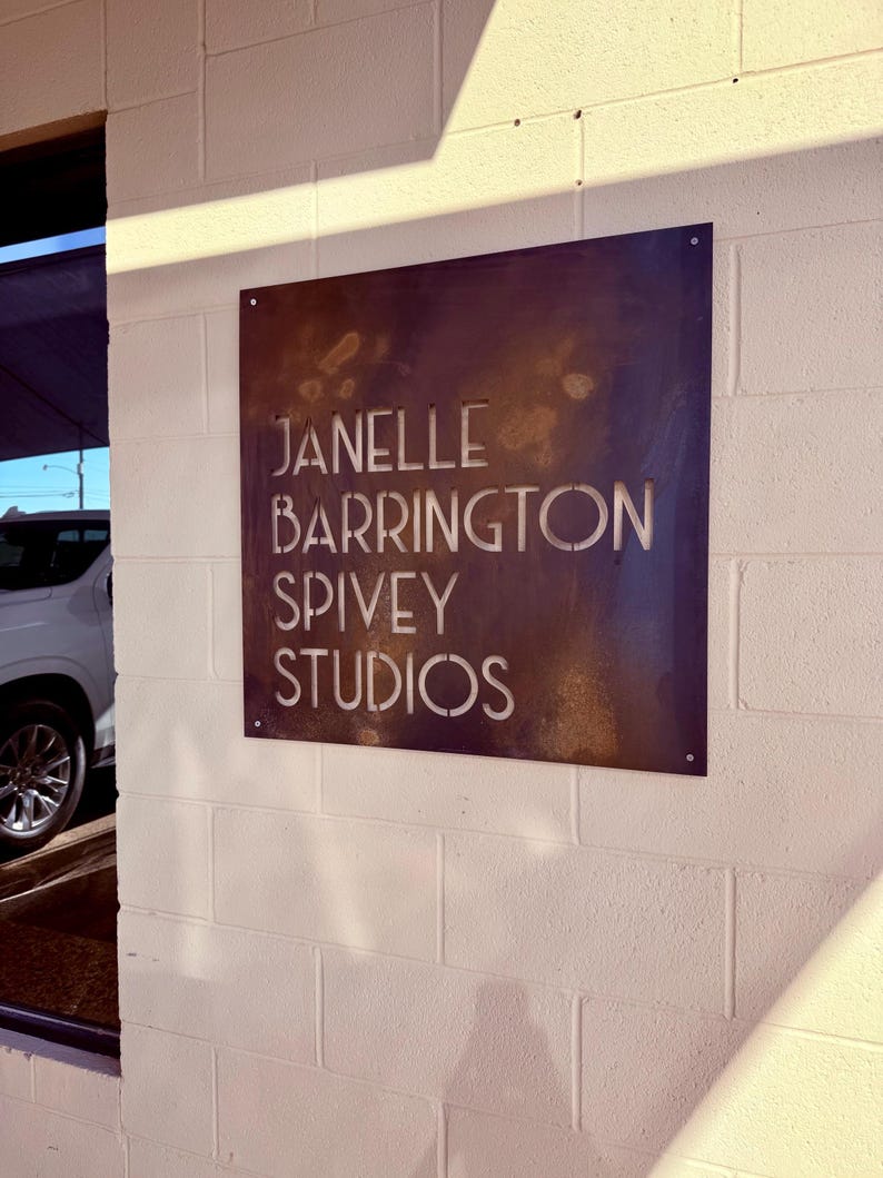 May include: A square, weathered metal sign with the text "JANELLE BARRINGTON SPIVEY STUDIOS" cut out. The sign is mounted on a white brick wall. The sign has a dark brown, rust-like appearance.