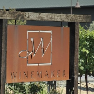May include: A rustic sign for a winery, featuring a weathered brown metal panel with the word "WINEMAKER" and a stylized logo. The sign is mounted on wooden posts, with a vineyard in the background.