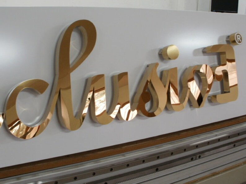 Puede incluir: Letras tridimensionales de color dorado que deletrean "lusion" con un s&iacute;mbolo de marca registrada. Las letras est&aacute;n montadas en una superficie blanca.