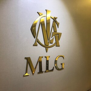 以下が含まれることがあります： 「MLG」という文字と、その上に「NA」という文字のスタイリッシュなモノグラムが書かれた金色のロゴです。
