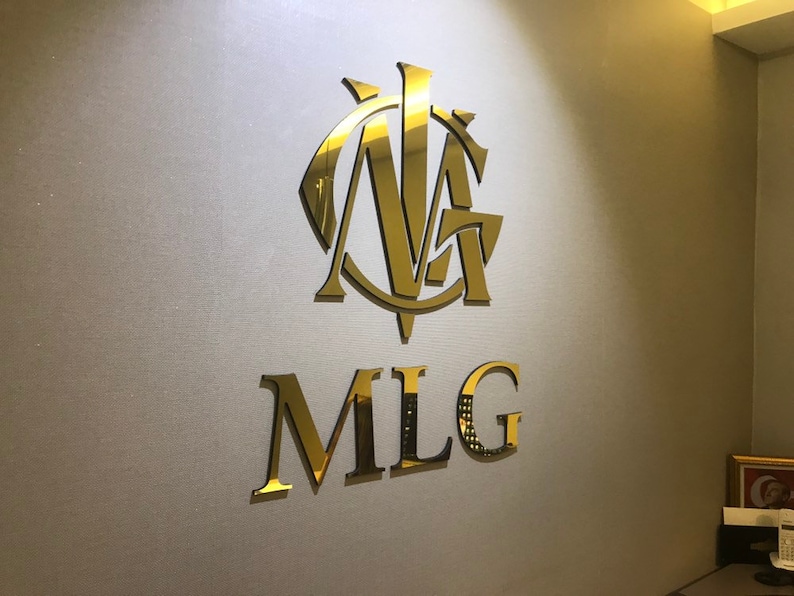 以下が含まれることがあります： 「MLG」という文字と、その上に「NA」という文字のスタイリッシュなモノグラムが書かれた金色のロゴです。