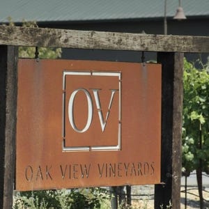 Op de afbeelding: Een rechthoekig bord van roestig metaal dat aan een donkerhouten frame hangt. Het bord heeft een vierkante uitsparing in het midden met de letters OV en de tekst OAK VIEW VINEYARDS in het verweerde metalen oppervlak gelaserd.