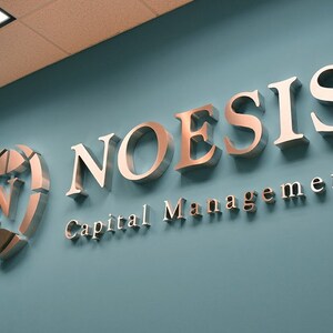 Puede incluir: Un letrero de metal plateado con el texto "NOESIS Capital Management" en una pared azul claro.