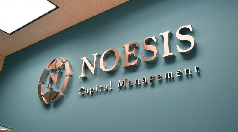 Puede incluir: Un letrero de metal plateado con el texto "NOESIS Capital Management" en una pared azul claro.