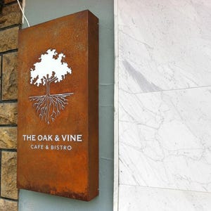 Op de afbeelding: Een rechthoekig, roestkleurig metalen bord gemonteerd aan een muur. Het bord bevat een witte uitgesneden afbeelding van een eikenboom met zichtbare wortels. Onder de boom staat de tekst THE OAK & VINE CAFE & BISTRO in witte hoofdletters.
