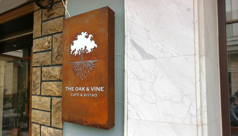 Op de afbeelding: Een rechthoekig, roestkleurig metalen bord gemonteerd aan een muur. Het bord bevat een witte uitgesneden afbeelding van een eikenboom met zichtbare wortels. Onder de boom staat de tekst THE OAK & VINE CAFE & BISTRO in witte hoofdletters.