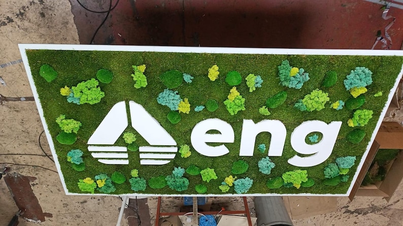 May include: A rectangular wall sign featuring a white logo and the lowercase text "eng" centered on a base of preserved green moss. The moss surface is decorated with various shades of green and light blue lichen clusters in different sizes.