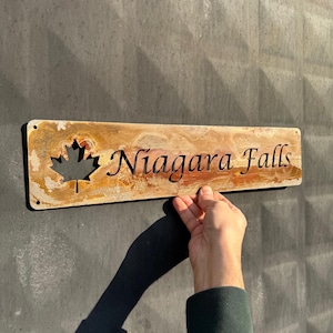 May include: A rectangular metal sign with the words "Niagara Falls" cut out in a decorative font. A maple leaf is cut out on the left side. The sign has a rustic, weathered appearance with shades of brown and gold.