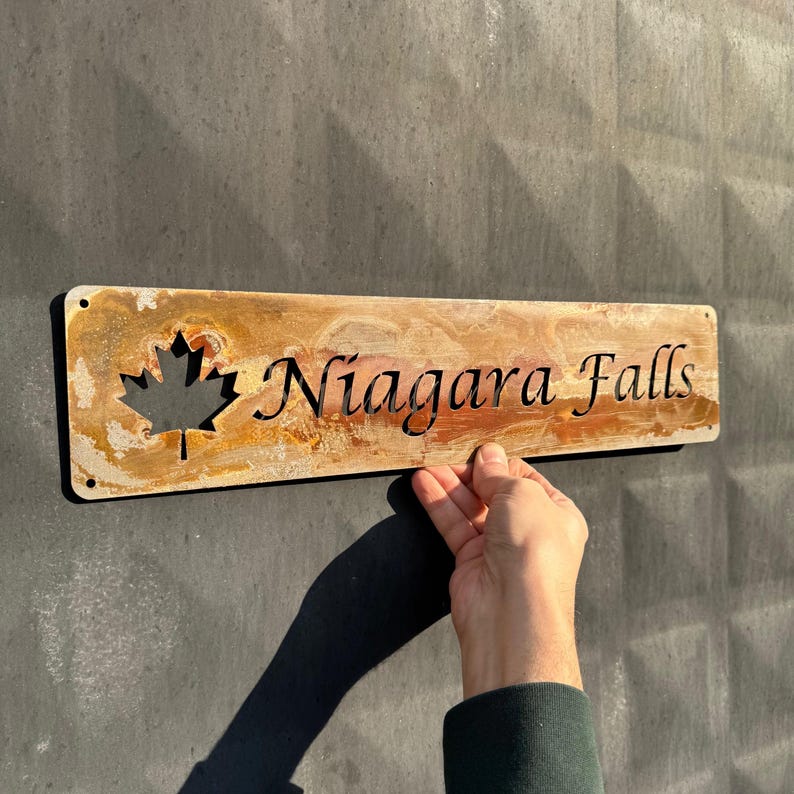 May include: A rectangular metal sign with the words "Niagara Falls" cut out in a decorative font. A maple leaf is cut out on the left side. The sign has a rustic, weathered appearance with shades of brown and gold.