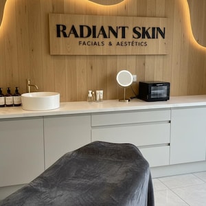May include: A treatment room featuring a white countertop with a sink, a gold faucet, skincare bottles, a round lighted mirror, and a black towel warmer. A wooden wall panel displays the black text RADIANT SKIN FACIALS & AEST&Eacute;TICS with backlighting.