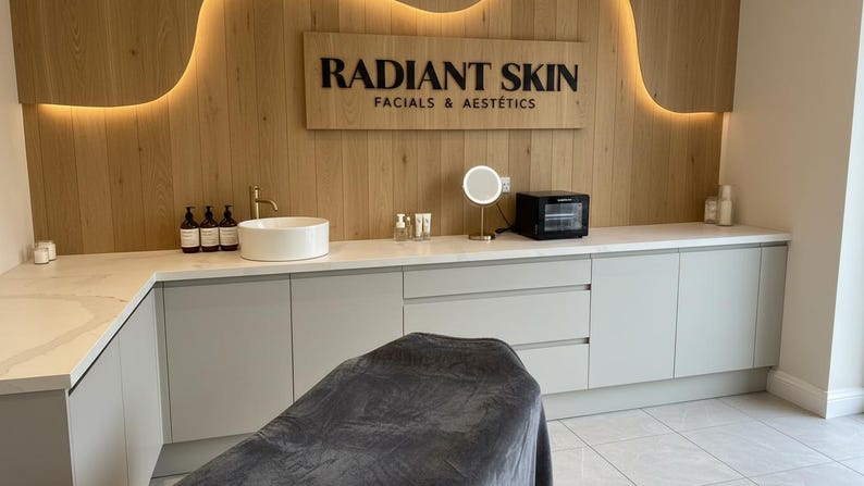 May include: A treatment room featuring a white countertop with a sink, a gold faucet, skincare bottles, a round lighted mirror, and a black towel warmer. A wooden wall panel displays the black text RADIANT SKIN FACIALS & AEST&Eacute;TICS with backlighting.