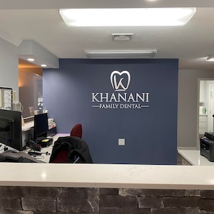 Puede incluir: Una sala de espera de un consultorio dental con un mostrador de recepci&oacute;n, dos computadoras, una silla y un letrero en la pared que dice "Khanani - Family Dental".