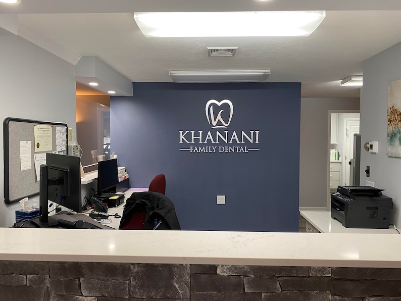 Puede incluir: Una sala de espera de un consultorio dental con un mostrador de recepci&oacute;n, dos computadoras, una silla y un letrero en la pared que dice "Khanani - Family Dental".