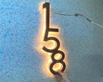 May include: Black illuminated house numbers 158 mounted on a wall. The numbers are lit from behind with a warm white light.