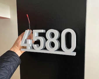 May include: A set of four silver-colored, three-dimensional house numbers, spelling out the number 4580. The numbers are attached to a black surface.