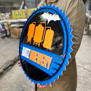 May include: A circular sign with a blue serrated edge resembling a bottle cap. The center features a black background with three orange brewing tanks and a white bat silhouette above. The text BATZ BREWERY is displayed in white and orange letters.