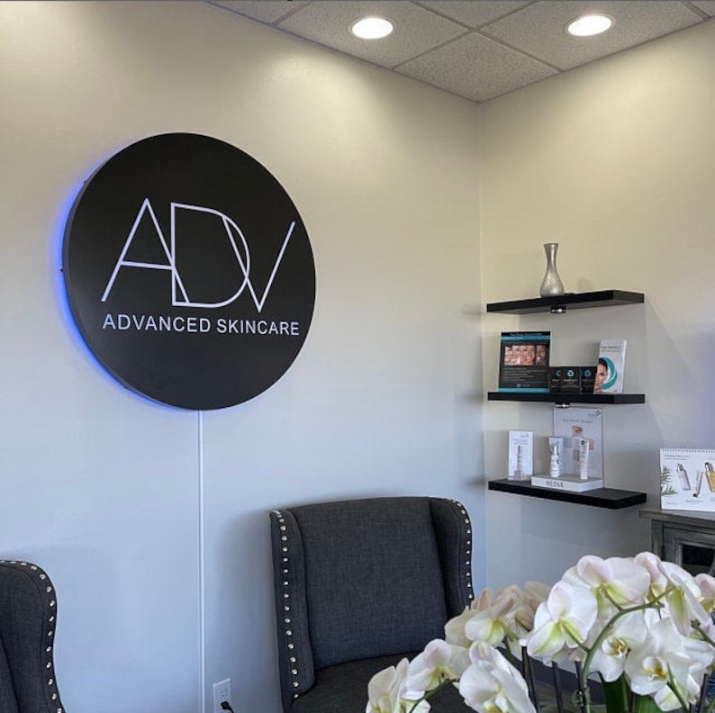 May include: A black circular sign with white lettering that reads "ADV ADVANCED SKINCARE". The sign is illuminated with a blue light. Two black chairs with gray upholstery are in the foreground. A white vase with a flower arrangement is on a shelf in the background.
