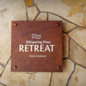 May include: A square, brown sign with the words "Whispering Pines RETREAT" in white lettering, mounted on a stone wall. The sign also includes the text "PRIVATE RESIDENCES" and is secured with four brass-colored rivets.
