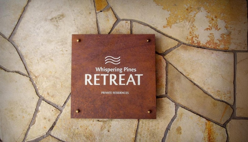 May include: A square, brown sign with the words "Whispering Pines RETREAT" in white lettering, mounted on a stone wall. The sign also includes the text "PRIVATE RESIDENCES" and is secured with four brass-colored rivets.