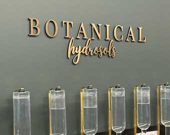 Könnte beinhalten: Eine Anzeige mit transparenten Glasspendern, die mit Flüssigkeit gefüllt sind, vor einer gedämpften grünen Wand. Die Worte "BOTANICAL hydrosols" stehen in goldfarbenen Buchstaben über den Spendern. Die Spender haben goldfarbene Metallakzente.