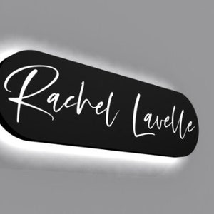 May include: Black oval sign with white script lettering that reads "Rachel Lavelle". The sign is illuminated with a white halo around the edges.