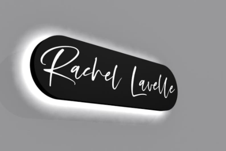 May include: Black oval sign with white script lettering that reads "Rachel Lavelle". The sign is illuminated with a white halo around the edges.