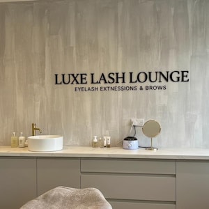 May include: A salon interior featuring a light gray textured wall with black lettering that reads LUXE LASH LOUNGE EYELASH EXTNESSIONS & BROWS. A white countertop holds a white basin, a gold faucet, various bottles, and a small round mirror.