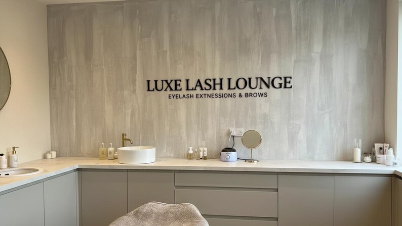 May include: A salon interior featuring a light gray textured wall with black lettering that reads LUXE LASH LOUNGE EYELASH EXTNESSIONS & BROWS. A white countertop holds a white basin, a gold faucet, various bottles, and a small round mirror.