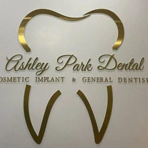 May include: A gold-colored wall sign featuring a stylized tooth outline. The text reads Ashley Park Dental in a cursive font, with the words COSMETIC IMPLANT & GENERAL DENTISRTY printed in a sans-serif font below the main name.