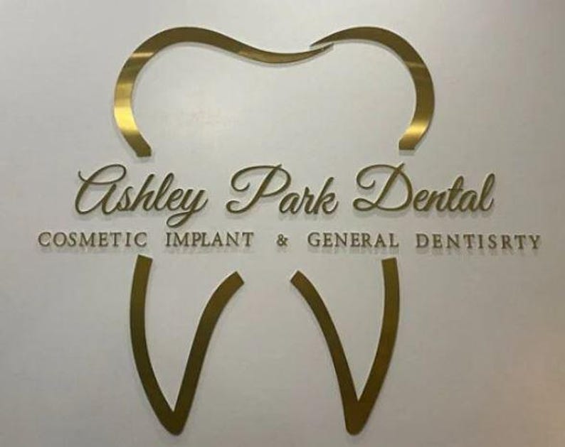 May include: A gold-colored wall sign featuring a stylized tooth outline. The text reads Ashley Park Dental in a cursive font, with the words COSMETIC IMPLANT & GENERAL DENTISRTY printed in a sans-serif font below the main name.