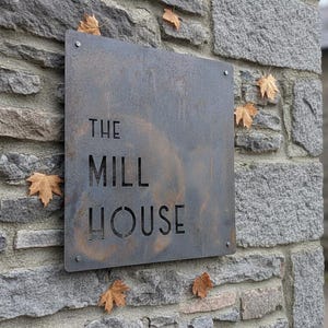 Op de afbeelding: Een vierkant, donker metalen bord met een roestige afwerking, bevestigd aan een grijze stenen muur. Het bord bevat de lasergesneden tekst "THE MILL HOUSE" in een strak, schreefloos lettertype. Vier kleine metalen bevestigingen houden de hoeken vast.