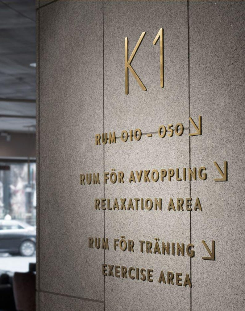 K&ouml;nnte beinhalten: Nahaufnahme einer Wand mit goldfarbenen Buchstaben und Richtungspfeilen. Der Text enth&auml;lt "K1", "Raum 010-050", "Entspannungsbereich" und "Fitnessbereich". Die Wand hat eine strukturierte, graue Oberfl&auml;che.