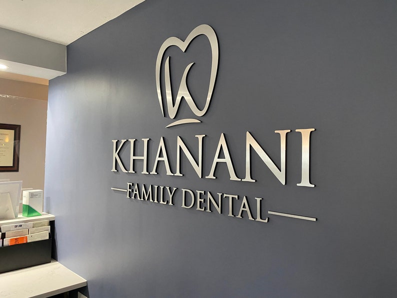 May include: A three-dimensional silver-colored wall sign featuring the text KHANANI FAMILY DENTAL in capital letters. Above the text is a stylized tooth icon containing the letter K. The sign is mounted on a solid dark gray wall surface.
