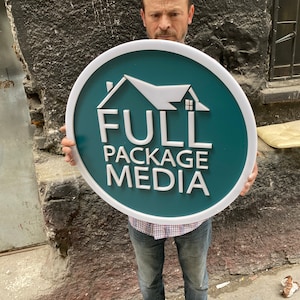 以下が含まれることがあります： 青緑色の背景に白い縁取りのある丸い看板を持った人がいます。看板には白い家のアイコンと白い文字で「FULL PACKAGE MEDIA」と書かれています。