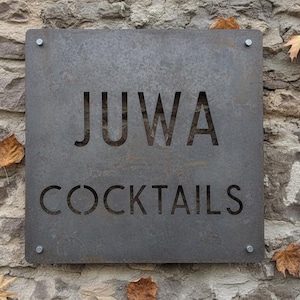 May include: A square, weathered metal sign with the word "JUWA" and the word "COCKTAILS" cut out of the metal. The sign is mounted on a rough stone wall, with a few fallen leaves.