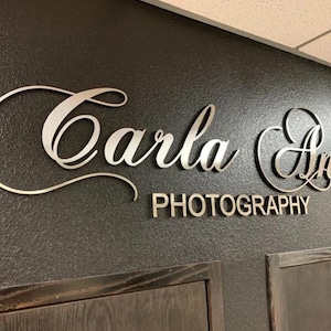 May include: A wall-mounted sign featuring the text Carla An in a large, silver-colored cursive script font, positioned above the word PHOTOGRAPHY in a smaller, silver-colored sans-serif font, all set against a dark, textured wall surface.