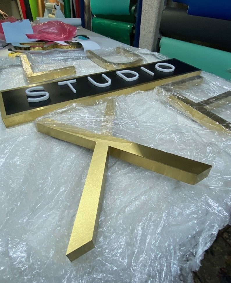 Puede incluir: Un letrero de metal de color dorado con la palabra "STUDIO" en letras blancas sobre fondo negro. El letrero est&aacute; envuelto en pl&aacute;stico y se encuentra sobre una superficie blanca.