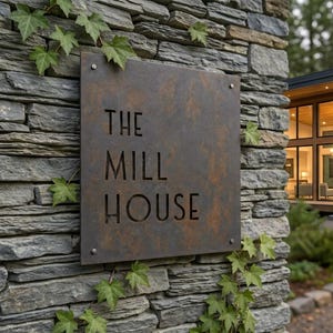 Könnte beinhalten: Ein quadratisches, verwittertes Metallschild mit dem Schriftzug THE MILL HOUSE in einer serifenlosen Schriftart, montiert an einer grauen Natursteinmauer mit grünen Efeublättern, die um die Ränder der Metallplatte und den Stein wachsen.