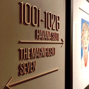 K&ouml;nnte beinhalten: Ein Wandschild mit dem Text "1001-1026 Havana Suite" und "The Magnificent Seven" in Hellbraun. Das Schild enth&auml;lt Richtungspfeile. Auf der rechten Seite des Bildes ist ein gerahmtes Portr&auml;t zu sehen.