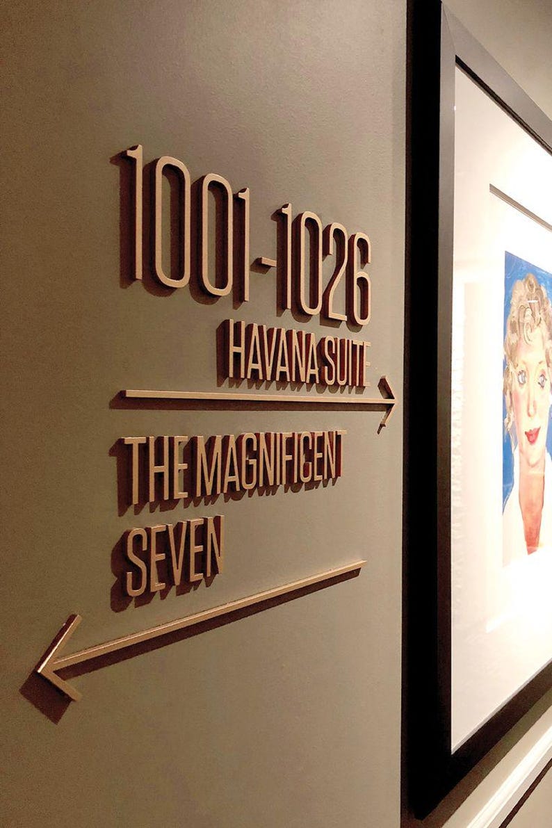K&ouml;nnte beinhalten: Ein Wandschild mit dem Text "1001-1026 Havana Suite" und "The Magnificent Seven" in Hellbraun. Das Schild enth&auml;lt Richtungspfeile. Auf der rechten Seite des Bildes ist ein gerahmtes Portr&auml;t zu sehen.
