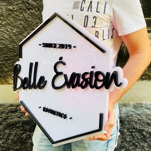 May include: A hexagonal white sign with a black border and raised black lettering that reads Belle &Eacute;vasion, with the text Since 2019 above and Esthetics below. The sign features a clean, layered design with bold, dark typography on a white base.