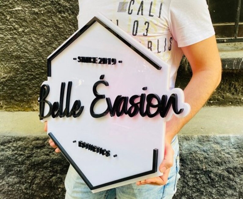 May include: A hexagonal white sign with a black border and raised black lettering that reads Belle &Eacute;vasion, with the text Since 2019 above and Esthetics below. The sign features a clean, layered design with bold, dark typography on a white base.