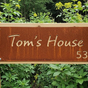 May include: A wooden sign with the text "Tom's House 53" cut out of a rusty brown metal plate. The sign is mounted on two wooden posts and surrounded by green foliage.