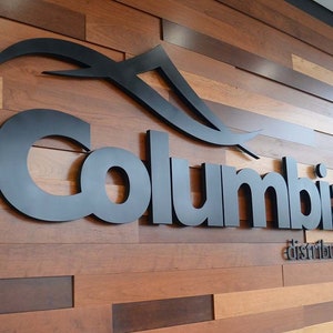 May include: A three-dimensional black sign featuring the word Columbia in a bold, sans-serif font with a stylized mountain graphic above it and the word distributing in a smaller font below, mounted on a wall of horizontal wood planks.