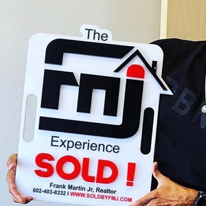 May include: A white rectangular sign with a handle cutout featuring the black text 'The' above a large black 'fmj' logo with a red roof icon. Below, the text reads 'Experience', 'SOLD!' in red, 'Frank Martin Jr, Realtor', and '602-403-8332 | WWW.SOLDBYFMJ.COM'.
